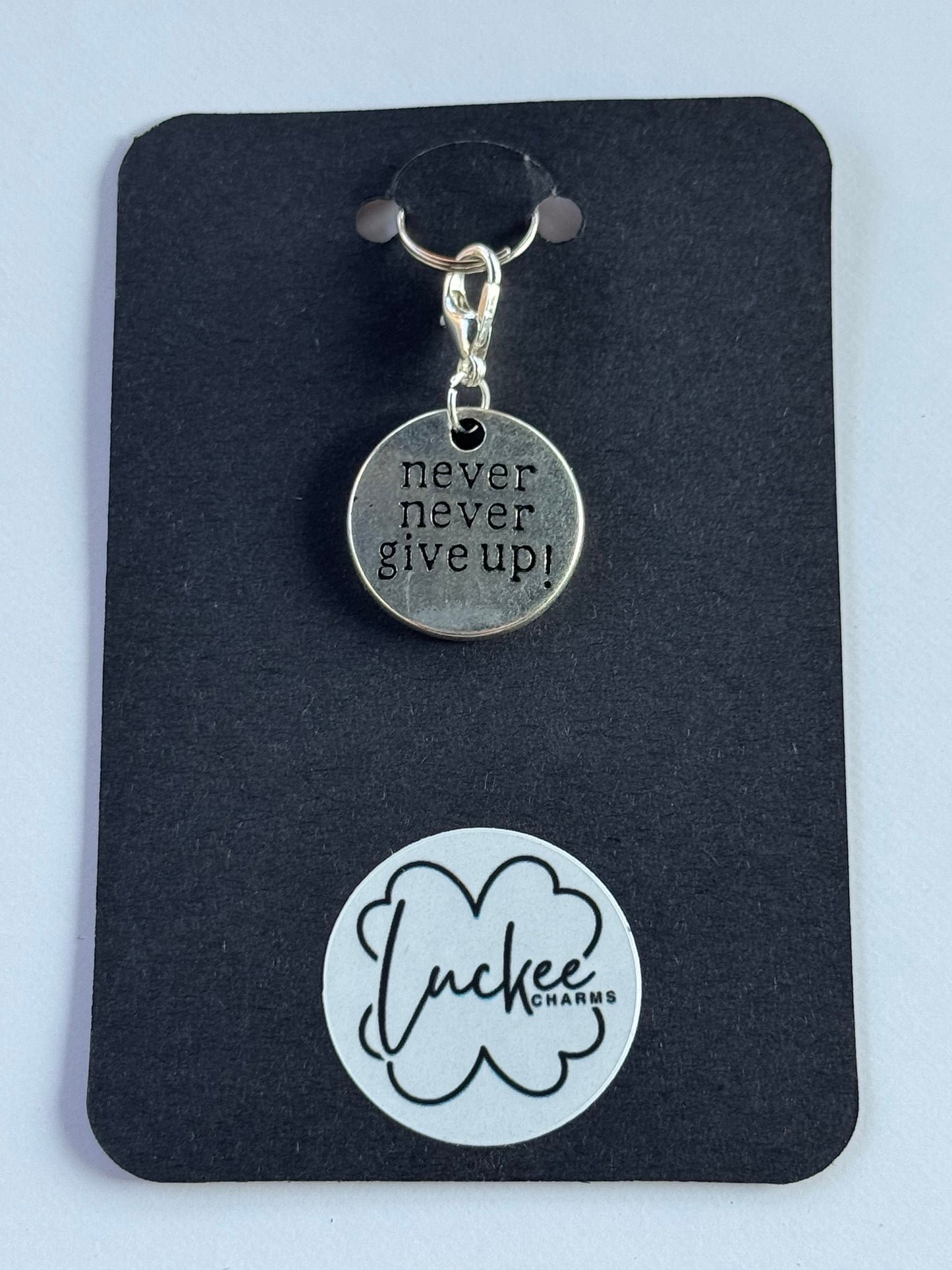 Silver charm with 'Never give up!' on a black card with a brand logo.