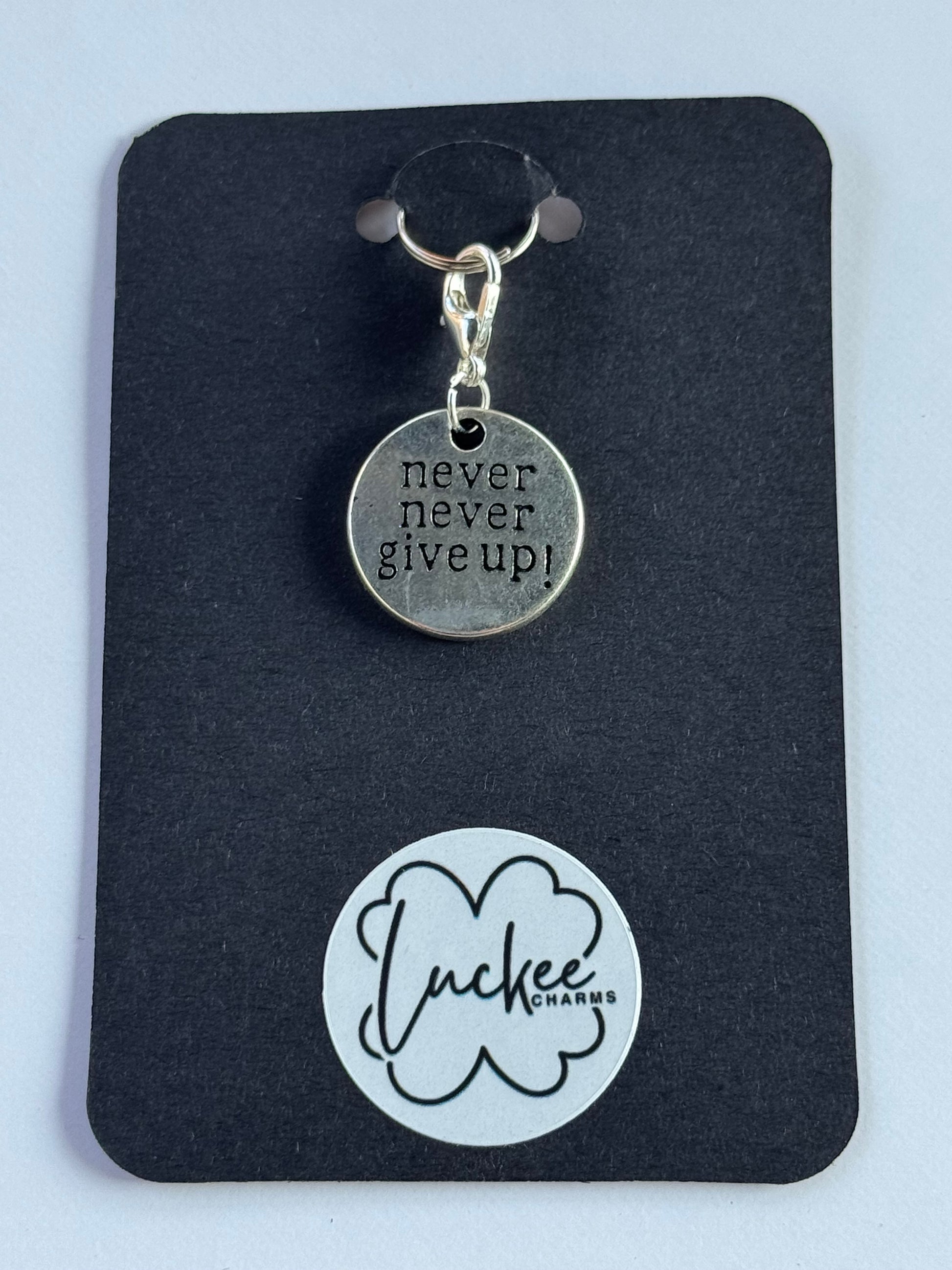 Silver charm with 'Never give up!' on a black card with a brand logo.