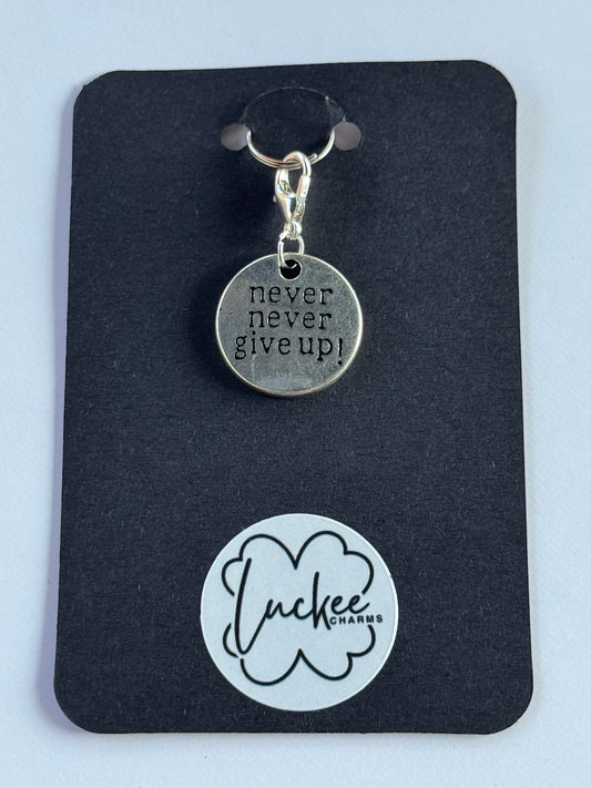 Silver charm with 'Never give up!' on a black card with a brand logo.